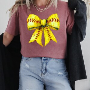 K&ouml;nnte beinhalten: Ein mauvefarbenes T-Shirt mit einem gelben und roten Softball-Schleifendesign. Die Schleife ist so gestaltet, dass sie einer Softball mit roten N&auml;hten &auml;hnelt.