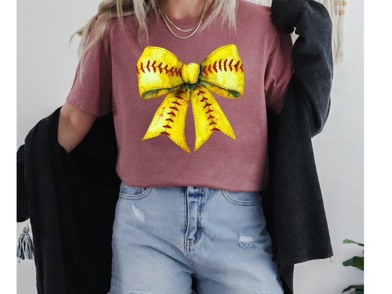 K&ouml;nnte beinhalten: Ein mauvefarbenes T-Shirt mit einem gelben und roten Softball-Schleifendesign. Die Schleife ist so gestaltet, dass sie einer Softball mit roten N&auml;hten &auml;hnelt.
