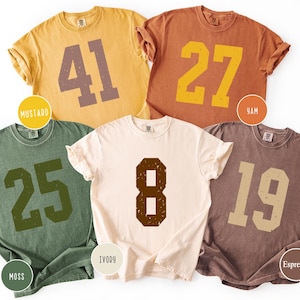 May include: Five short-sleeved t-shirts in mustard, yam, moss, ivory, and espresso colours. Each shirt features a large, distressed number in a contrasting colour. The shirts are casual wear.