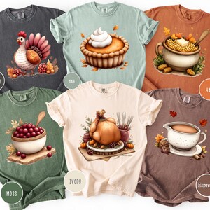 May include: A collection of six t-shirts in various colors, each featuring a watercolor illustration of Thanksgiving-themed food items. Designs include a turkey, pie, and cranberry sauce. The shirts are labeled with color names like "Pepper" and "Ivory".