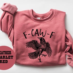 May include: Heather scarlet red sweatshirt with the text "F-CAW-F" and a black raven illustration. The raven is depicted with outstretched wings and a detailed, artistic design.