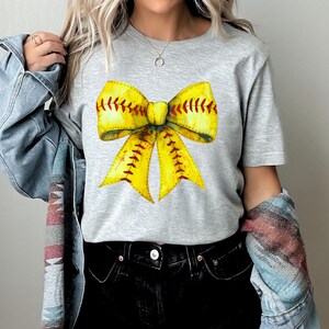 K&ouml;nnte beinhalten: Ein graues T-Shirt mit einem gelben und roten Softball-Schleifendesign. Die Schleife ist so gestaltet, dass sie einem Softball mit roten N&auml;hten &auml;hnelt.