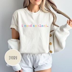 May include: Ivory-colored t-shirt with the text "i need an alani" in rainbow colors. The shirt is part of a casual outfit, paired with gray shorts and a white cardigan. The overall style is relaxed and comfortable.