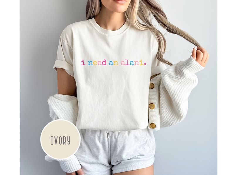 May include: Ivory-colored t-shirt with the text "i need an alani" in rainbow colors. The shirt is part of a casual outfit, paired with gray shorts and a white cardigan. The overall style is relaxed and comfortable.