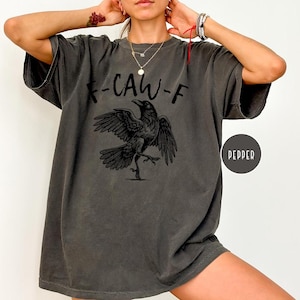 Peut inclure: T-shirt gris fonc&eacute; avec un motif de corbeau noir et le texte "-CAW-F". Le corbeau est repr&eacute;sent&eacute; avec les ailes d&eacute;ploy&eacute;es et une patte lev&eacute;e. Le t-shirt a une &eacute;tiquette de couleur poivre.