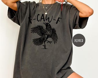 Könnte beinhalten: Dunkelgraues T-Shirt mit einer schwarzen Raben-Grafik und dem Text "-CAW-F". Der Rabe wird mit ausgebreiteten Flügeln und einem erhobenen Bein dargestellt. Das Shirt hat ein Pepper-Farbetikett.