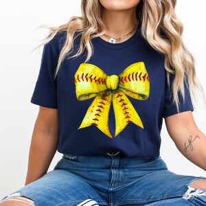 K&ouml;nnte beinhalten: Ein marineblaues T-Shirt mit einem gelben Softball-Schleifendesign. Die Schleife besteht aus einem gelben Softball mit roten N&auml;hten.