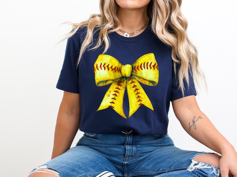 K&ouml;nnte beinhalten: Ein marineblaues T-Shirt mit einem gelben Softball-Schleifendesign. Die Schleife besteht aus einem gelben Softball mit roten N&auml;hten.