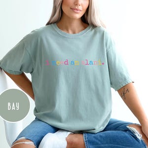 May include: A light green t-shirt with the text "I need a land" in rainbow colors. The shirt is a crew neck style and is worn with blue jeans. The shirt is a relaxed fit.