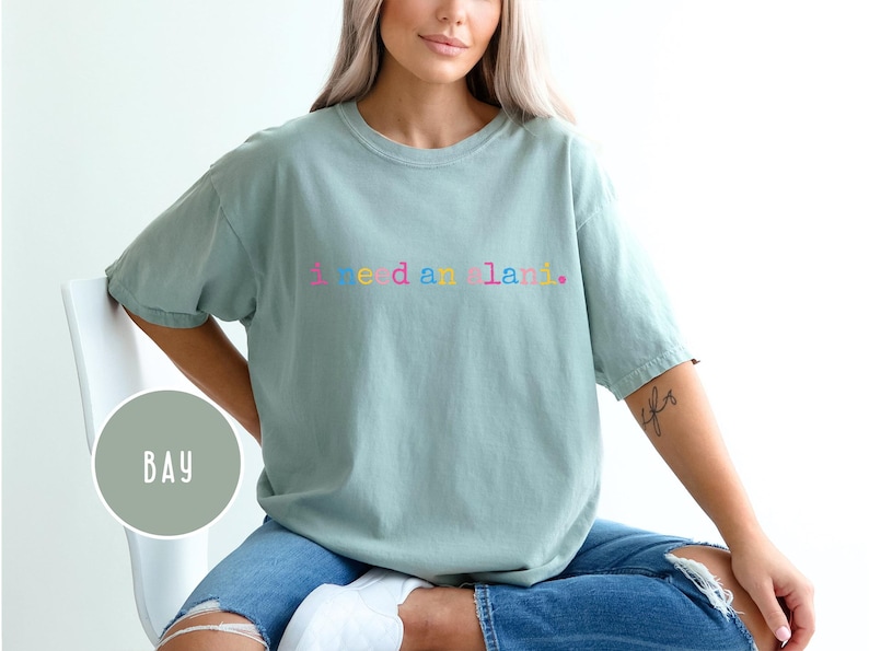 May include: A light green t-shirt with the text "I need a land" in rainbow colors. The shirt is a crew neck style and is worn with blue jeans. The shirt is a relaxed fit.