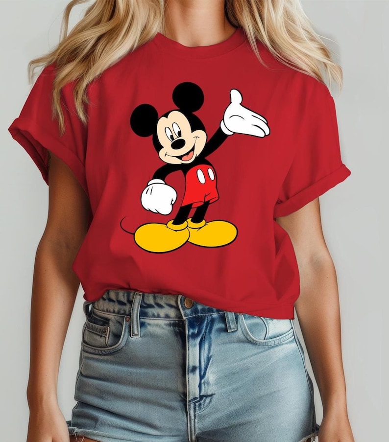 Puede incluir: Camiseta roja con un dibujo de Mickey Mouse. El personaje de dibujos animados tiene orejas negras, cuerpo negro, pantalones cortos rojos, zapatos amarillos y guantes blancos. La camiseta tiene mangas enrolladas.