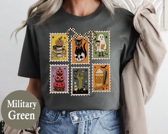 May include: Military green t-shirt featuring a Halloween-themed stamp design. The stamps depict a pumpkin, black cat, ghost, stacked pumpkins, zombie hand, and a coffee cup. The design is accented with a checkered bow.