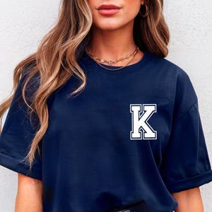May include: Navy blue t-shirt featuring a white and navy letter "K" on the front. The short-sleeved shirt has a crew neck and a relaxed fit. The shirt is made of a soft material.