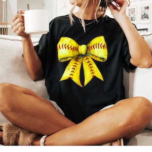 K&ouml;nnte beinhalten: Ein schwarzes T-Shirt mit einem gelben und roten Softball-Schleifendesign. Die Schleife ist so gestaltet, dass sie einer Softball mit roten N&auml;hten &auml;hnelt.