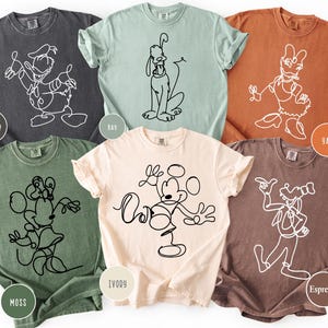 May include: Six different colored t-shirts, each featuring a unique line drawing of a cartoon character. The shirts are in shades of gray, green, light blue, orange, ivory, and brown. The characters include Donald Duck, Pluto, Daisy Duck, Minnie Mouse, Mickey Mouse, and Goofy.
