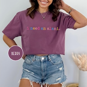 May include: Berry-colored t-shirt with the text "i need an alani" in rainbow-colored letters. The shirt is a short-sleeved, crew neck style. The word "BERRY" is in a circle on the bottom left of the image.