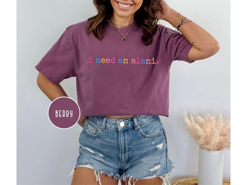 May include: Berry-colored t-shirt with the text "i need an alani" in rainbow-colored letters. The shirt is a short-sleeved, crew neck style. The word "BERRY" is in a circle on the bottom left of the image.