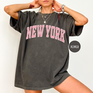 Peut inclure: T-shirt gris foncé surdimensionné avec l'inscription "NEW YORK" en rose. Le t-shirt a des manches courtes et une coupe ample. Un petit logo circulaire avec le mot "PEPPER" est visible.
