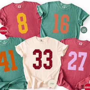 May include: Five short-sleeved t-shirts in shades of crimson, light green, and ivory. Each shirt displays a large number in a contrasting colour. Numbers include 8, 16, 41, 33, and 27. The shirts have a vintage, worn appearance.