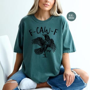 Peut inclure: T-shirt de couleur sarcelle avec le texte "F-CAW-F" et une illustration d'un corbeau noir. Le corbeau a les ailes d&eacute;ploy&eacute;es et une patte lev&eacute;e. Le t-shirt est de style d&eacute;contract&eacute;.
