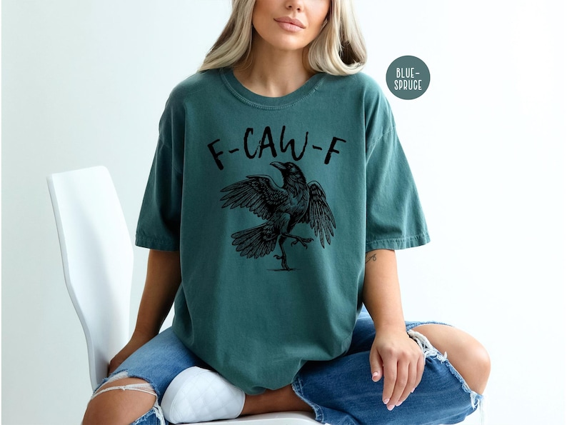 Peut inclure: T-shirt de couleur sarcelle avec le texte "F-CAW-F" et une illustration d'un corbeau noir. Le corbeau a les ailes d&eacute;ploy&eacute;es et une patte lev&eacute;e. Le t-shirt est de style d&eacute;contract&eacute;.