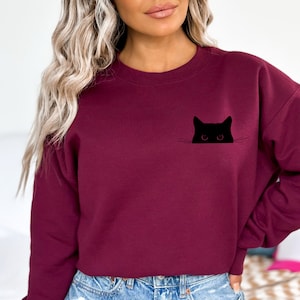 May include: Burgundy sweatshirt featuring a black cat graphic. The cat's head and eyes peek out from a pocket-like design. The sweatshirt is a solid color, with a crew neck and long sleeves. A casual, comfortable style.