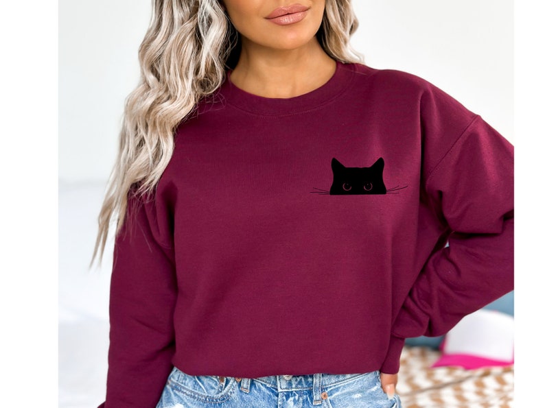 May include: Burgundy sweatshirt featuring a black cat graphic. The cat's head and eyes peek out from a pocket-like design. The sweatshirt is a solid color, with a crew neck and long sleeves. A casual, comfortable style.