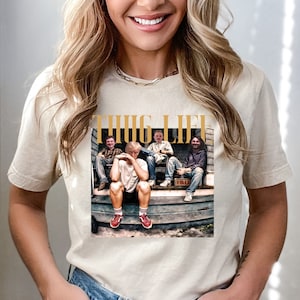 May include: A white t-shirt with a graphic print of four people sitting on a porch. The graphic is in a vintage style and features the text "THUG LIFE" in gold lettering.