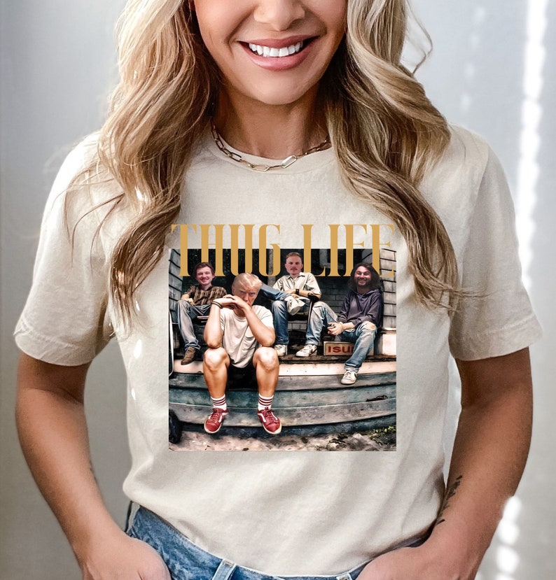 May include: A white t-shirt with a graphic print of four people sitting on a porch. The graphic is in a vintage style and features the text "THUG LIFE" in gold lettering.
