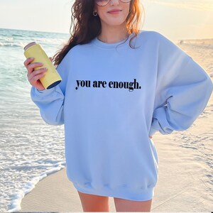 May include: A light blue sweatshirt with the text "you are enough." printed in black on the front. The sweatshirt is being worn by a person standing on a beach.