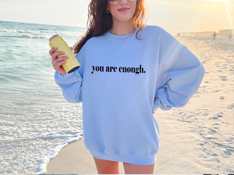 May include: A light blue sweatshirt with the text "you are enough." printed in black on the front. The sweatshirt is being worn by a person standing on a beach.