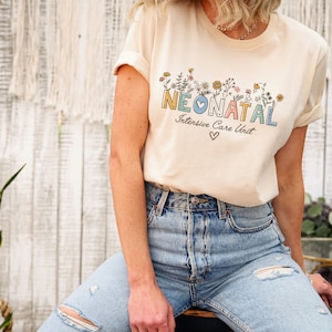 May include: A light beige t-shirt with a floral design and the text "NEONATAL Intensive Care Unit" in colorful letters. A small heart is below the text.