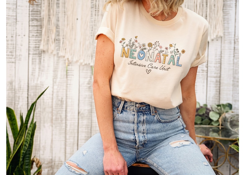 May include: A light beige t-shirt with a floral design and the text "NEONATAL Intensive Care Unit" in colorful letters. A small heart is below the text.
