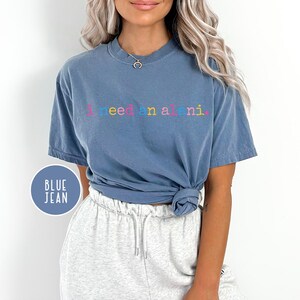 May include: Blue jean-colored t-shirt with the text "I need a mani" in colorful letters. The shirt is a short-sleeved, crew neck style. A blue circle with the words "BLUE JEAN" is in the lower left corner.