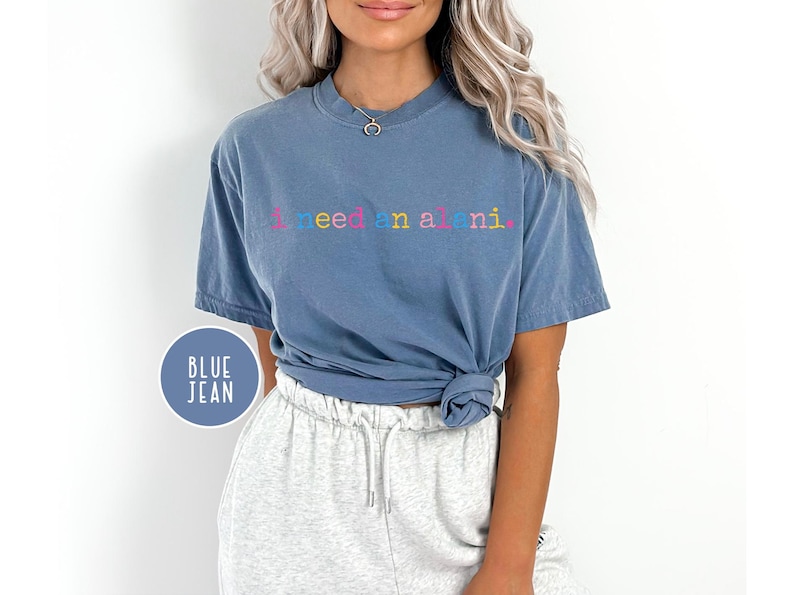May include: Blue jean-colored t-shirt with the text "I need a mani" in colorful letters. The shirt is a short-sleeved, crew neck style. A blue circle with the words "BLUE JEAN" is in the lower left corner.