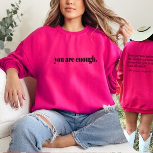 May include: A bright pink sweatshirt with the text "you are enough." printed on the front. The back of the sweatshirt has the text "Dear person behind me, the world is a better place with you in it. Lots of love." printed on it.