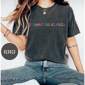 May include: A dark gray t-shirt with the text "i need an alani." in rainbow colors. The shirt is paired with blue jeans. The model is wearing a necklace and bracelets.