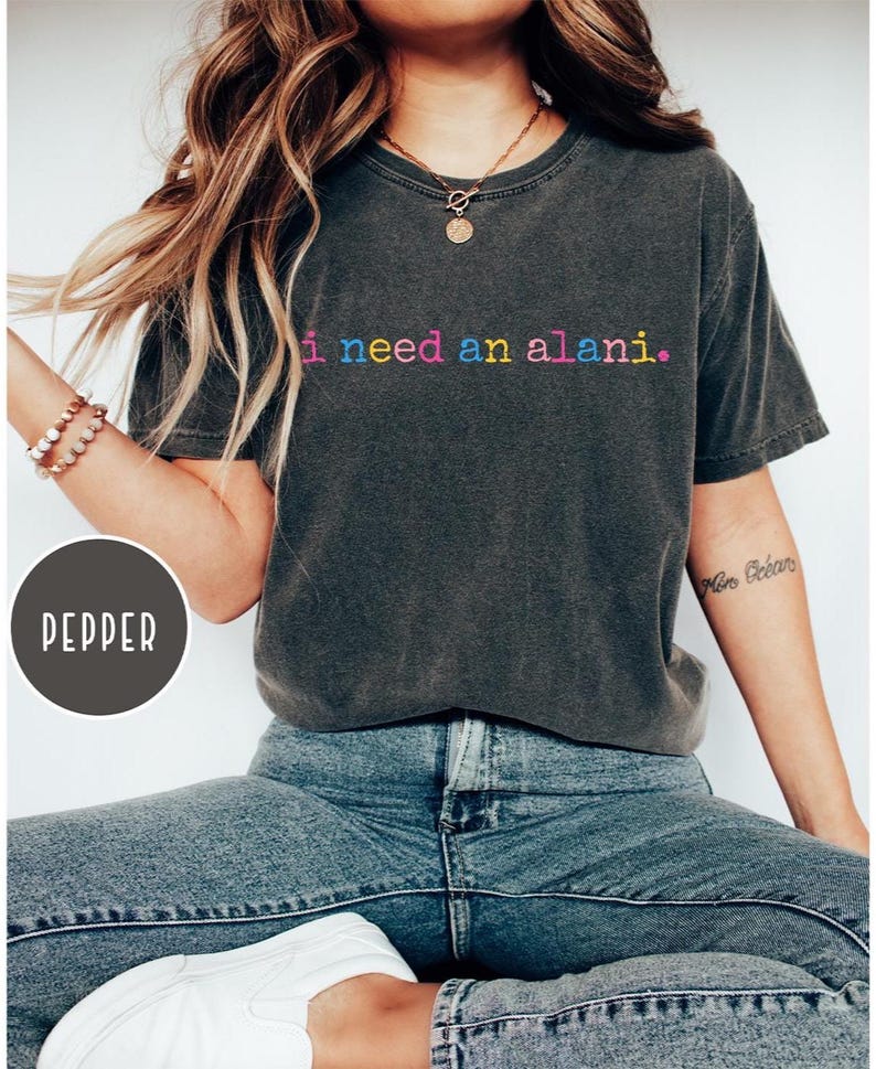 May include: A dark gray t-shirt with the text "i need an alani." in rainbow colors. The shirt is paired with blue jeans. The model is wearing a necklace and bracelets.