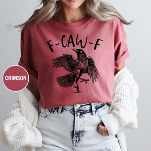 Peut inclure: T-shirt couleur pourpre avec l'inscription "F-CAW-F" et une illustration noire d'un corbeau aux ailes d&eacute;ploy&eacute;es. Le t-shirt est de couleur unie et a des manches retrouss&eacute;es.