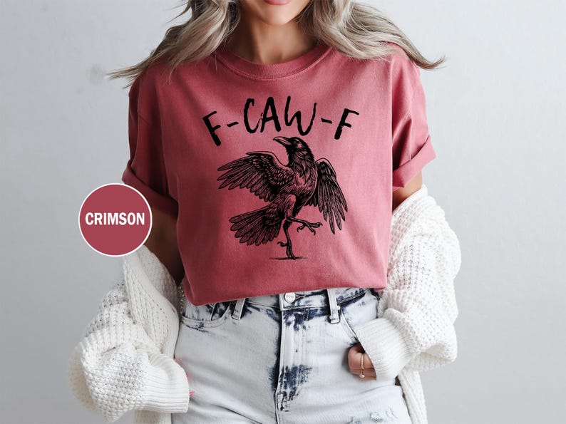 Peut inclure: T-shirt couleur pourpre avec l'inscription "F-CAW-F" et une illustration noire d'un corbeau aux ailes d&eacute;ploy&eacute;es. Le t-shirt est de couleur unie et a des manches retrouss&eacute;es.
