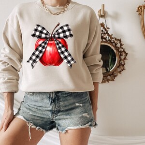 May include: A beige sweatshirt with a red apple and a black and white checkered bow. The apple has a green stem and leaf.