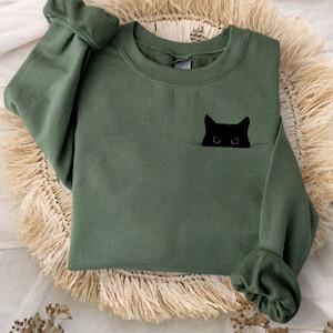 May include: Olive green sweatshirt with a black cat graphic. The cat's head is peeking out from the pocket area. The sweatshirt has long sleeves with cuffed wrists. The garment is displayed on a woven, fringed mat.