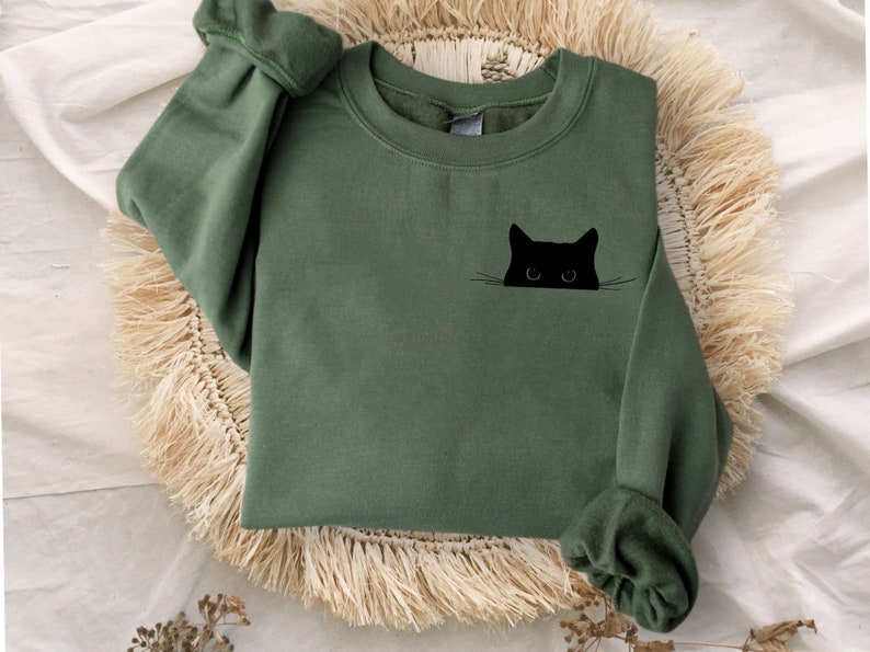 May include: Olive green sweatshirt with a black cat graphic. The cat's head is peeking out from the pocket area. The sweatshirt has long sleeves with cuffed wrists. The garment is displayed on a woven, fringed mat.