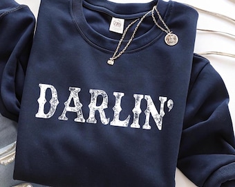Könnte beinhalten: Marineblaues Sweatshirt mit dem Wort "DARLIN'" in verblichenen weißen Buchstaben aufgedruckt. Eine goldene Halskette mit Anhänger liegt auf dem Sweatshirt. Das Sweatshirt ist auf einer weißen Oberfläche gefaltet.