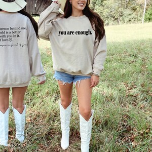May include: A beige sweatshirt with the text "you are enough." printed on the front. The back of the sweatshirt has the text "Dear person behind me, the world is a better place with you in it. Lots of love... - the person in front of you".