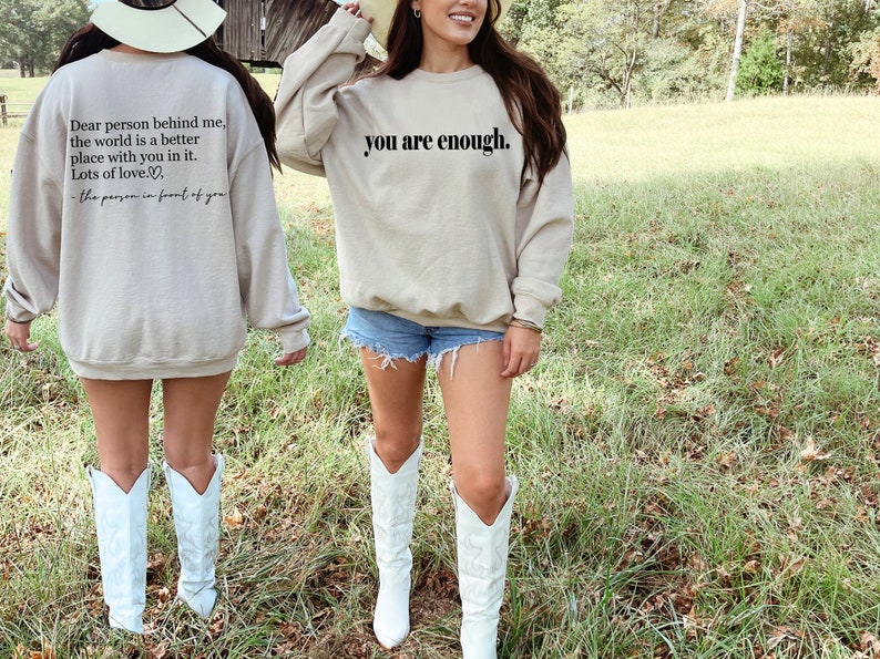 May include: A beige sweatshirt with the text "you are enough." printed on the front. The back of the sweatshirt has the text "Dear person behind me, the world is a better place with you in it. Lots of love... - the person in front of you".