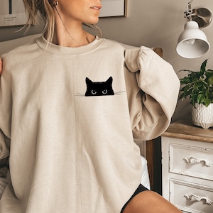 May include: Beige sweatshirt featuring a black cat graphic. The cat's head is peeking out from a pocket-like design. The sweatshirt has a crew neck and long sleeves. The overall style is casual and comfortable, suitable for everyday wear.