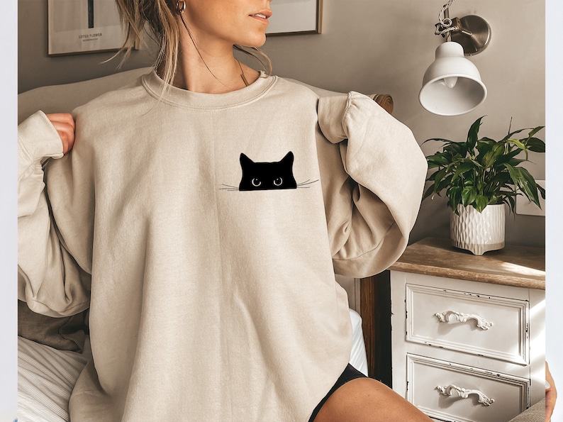 May include: Beige sweatshirt featuring a black cat graphic. The cat's head is peeking out from a pocket-like design. The sweatshirt has a crew neck and long sleeves. The overall style is casual and comfortable, suitable for everyday wear.