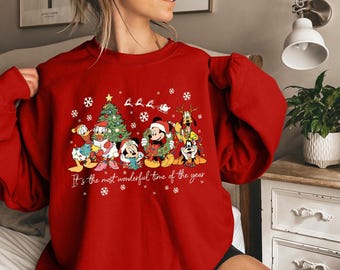 Könnte beinhalten: Rotes Sweatshirt mit einem weihnachtlichen Design mit Disney-Figuren wie Mickey Mouse und Donald Duck. Die Grafik enthält einen Weihnachtsbaum, Schneeflocken und den Text "It's the most wonderful time of the year".