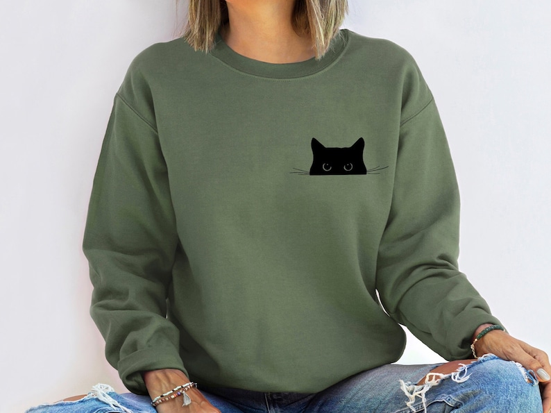 May include: Olive green sweatshirt featuring a black cat graphic. The cat's head and eyes peek over a line, creating a minimalist design. The sweatshirt is a solid color and has a crew neck.